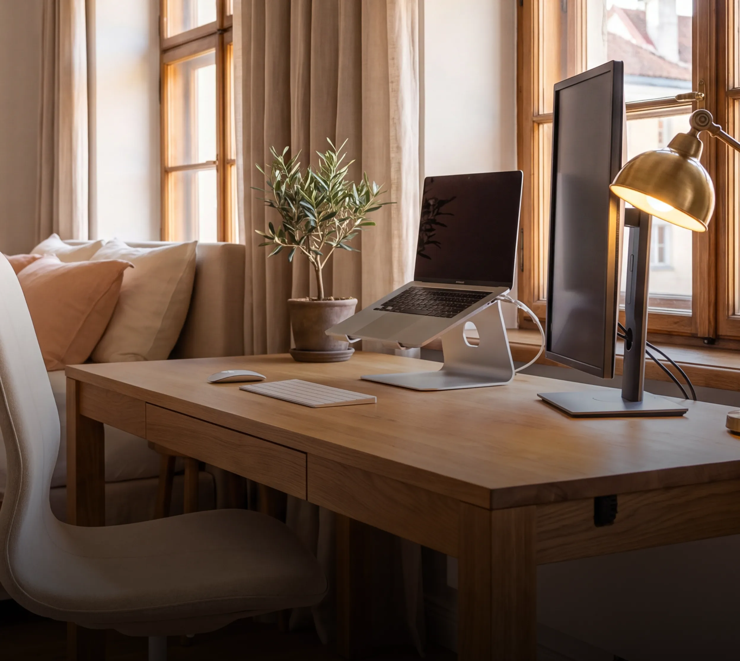 A cozy home desk setup with a laptop stand, monitor, and olive plant by a sunlit window