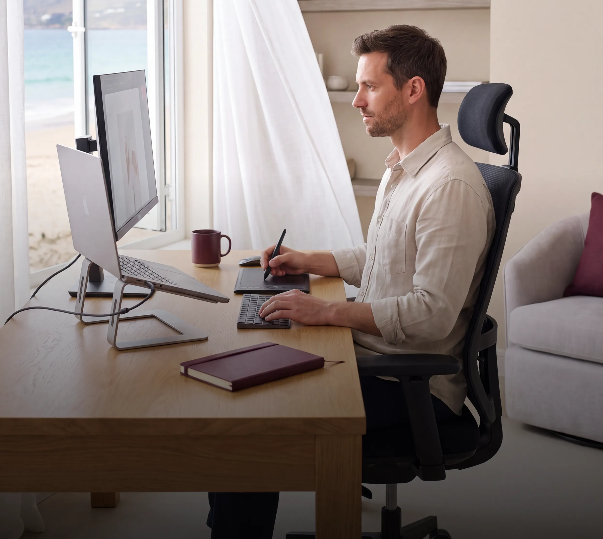 A man working at a standing desk with a beach and ocean visible through the window
